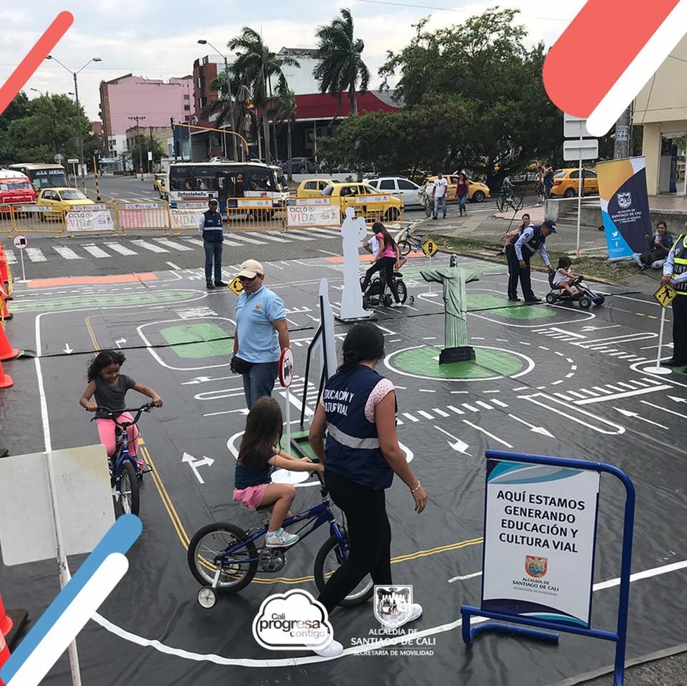 Niños en bicicletas y triciclos en una calle, siendo educados por personal de la Alcaldía sobre las normas de tránsito.